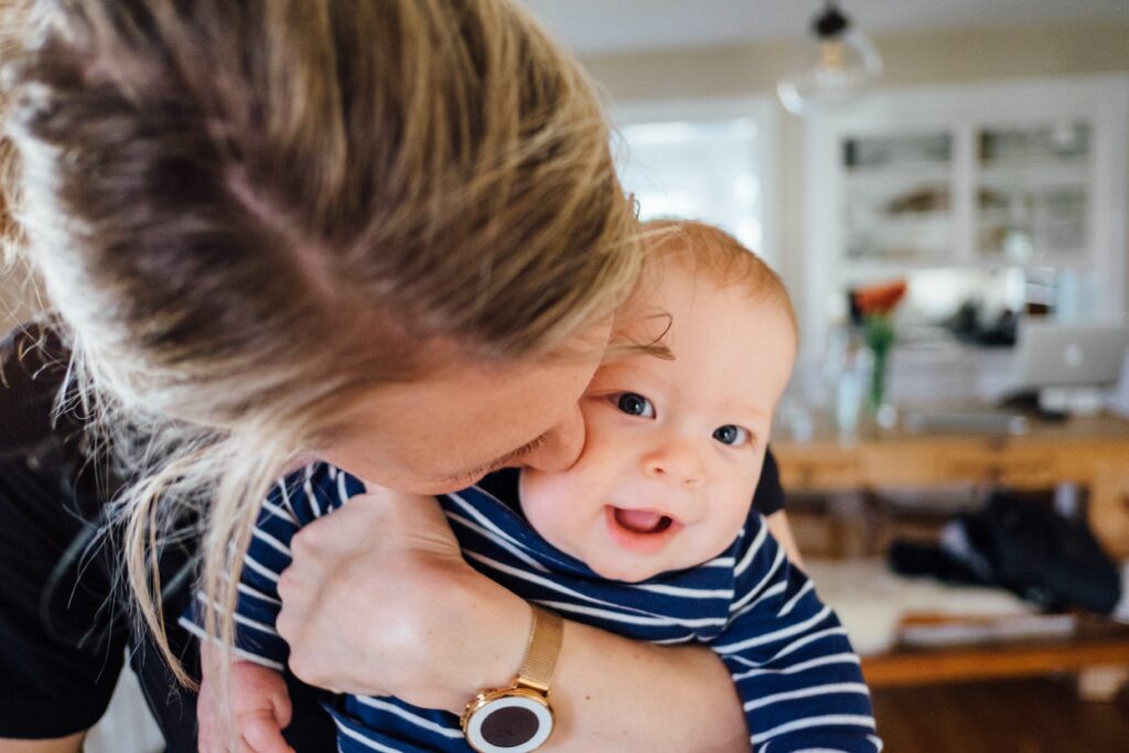 a white blonde lady stood close to the camera giving her baby a kiss on the head