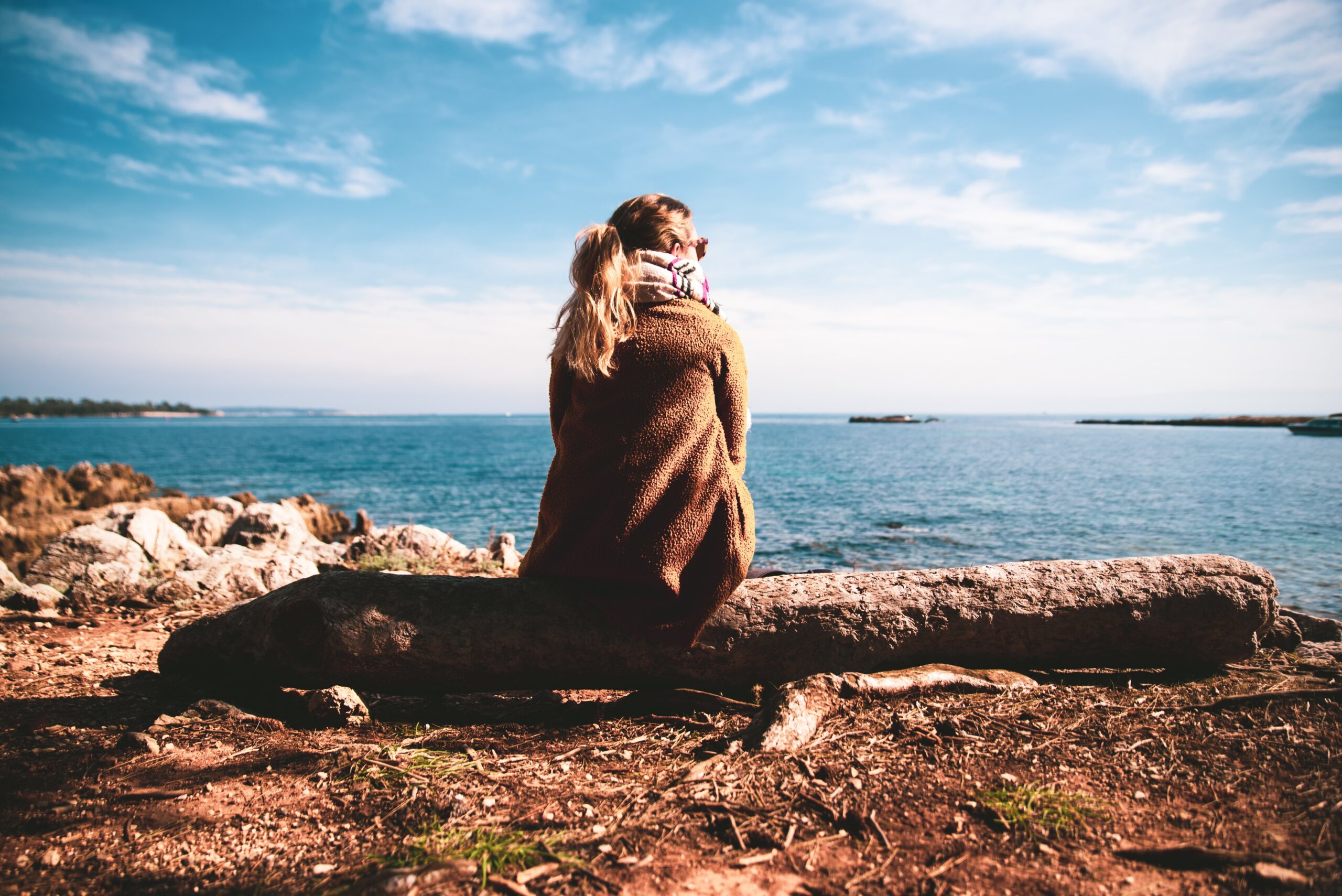 Girl in brown jacket and scarf sitting on log looking out to sea