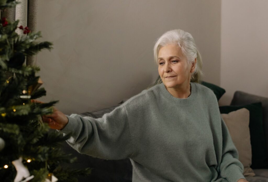 an older woman with white hair and a green jumper reaching out and touching a christmas tree