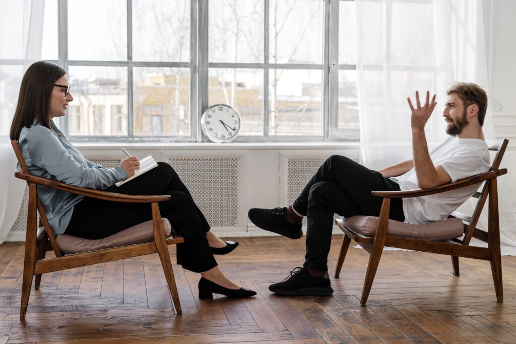 A woman and a man facing each other and talking in a room in front of a window